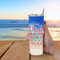Close-up of woman holding tumbler with beach in background and sunny, relaxed vibes.